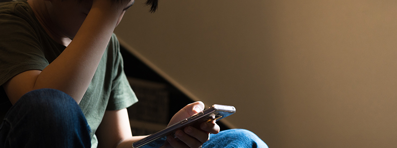 Young preteen/teenage boy sit on stair at home, cry and cover his face with hand, other hand hold smartphone, feeling alone, stressed, frustrated, overwhelmed, depressed. Cyber bullying concept.