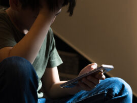 Young preteen/teenage boy sit on stair at home, cry and cover his face with hand, other hand hold smartphone, feeling alone, stressed, frustrated, overwhelmed, depressed. Cyber bullying concept.